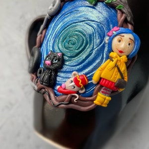 May include: A black mug with a blue and green swirl design. The mug is decorated with a small black cat, a mouse wearing a red and gold uniform, and a girl with red hair wearing a yellow jacket. The girl is holding a small black cat.