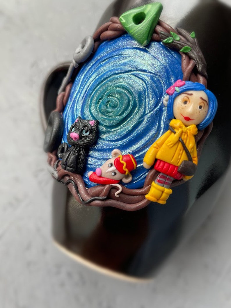 May include: A black mug with a blue and green swirl design. The mug is decorated with a small black cat, a mouse wearing a red and gold uniform, and a girl with red hair wearing a yellow jacket. The girl is holding a small black cat.