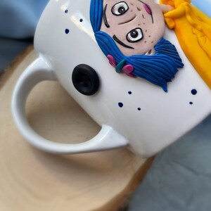 May include: White ceramic mug with a hand-painted illustration of a girl with blue hair and a yellow shirt. The girl has brown eyes and freckles. The mug has a black button detail and blue dots.