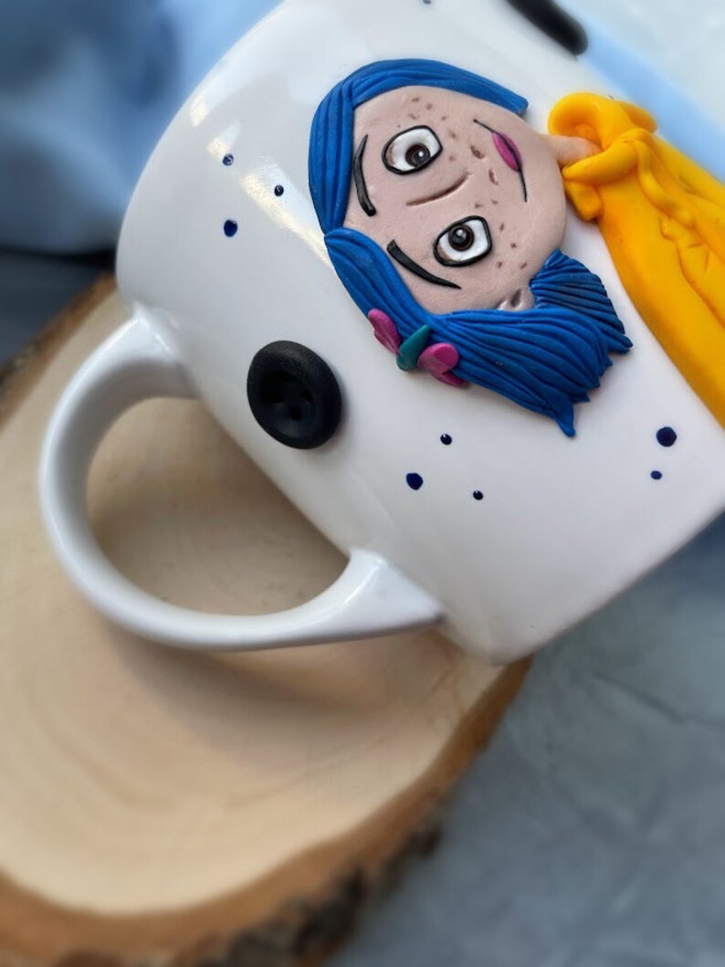 May include: White ceramic mug with a hand-painted illustration of a girl with blue hair and a yellow shirt. The girl has brown eyes and freckles. The mug has a black button detail and blue dots.