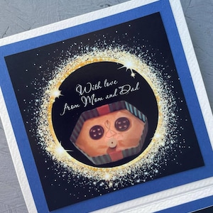 May include: A card with a black background and gold glitter. The card has a picture of a wrapped gift with a face and the text "With love from Mom and Dad".