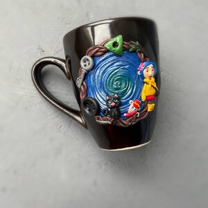 May include: A black ceramic mug with a blue and green swirl design. The mug has a 3D design of a girl with a yellow jacket, a black cat, and a mouse.