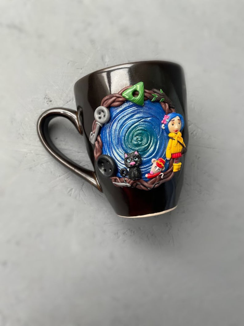 May include: A black ceramic mug with a blue and green swirl design. The mug has a 3D design of a girl with a yellow jacket, a black cat, and a mouse.