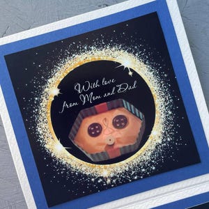 May include: A card with a black background and gold glitter. The card has a picture of a wrapped gift with a face and the text "With love from Mom and Dad".