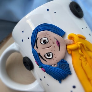 May include: White ceramic mug with a blue-haired character with yellow clothing sculpted on the front. The character has brown eyes and freckles. The mug has a black button on the handle.