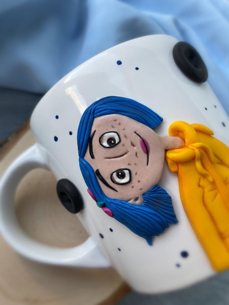 May include: White ceramic mug with a blue-haired character with yellow clothing sculpted on the front. The character has brown eyes and freckles. The mug has a black button on the handle.