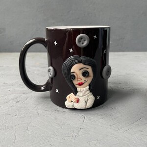 May include: A black ceramic mug with a white rim and handle. The mug features a 3D sculpted image of a woman with black hair, wearing a white sweater and red nail polish. The background is decorated with white stars and gray buttons.