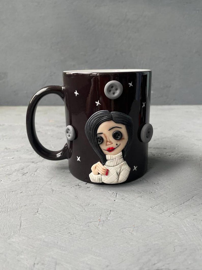 May include: A black ceramic mug with a white rim and handle. The mug features a 3D sculpted image of a woman with black hair, wearing a white sweater and red nail polish. The background is decorated with white stars and gray buttons.