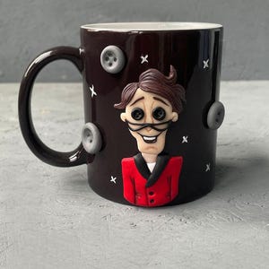 May include: Black ceramic mug with a 3D sculpted image of a character from the movie "The Nightmare Before Christmas". The character is wearing a red suit and has a white collar. The mug is decorated with gray buttons and white X's.