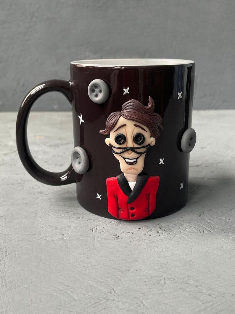 May include: Black ceramic mug with a 3D sculpted image of a character from the movie "The Nightmare Before Christmas". The character is wearing a red suit and has a white collar. The mug is decorated with gray buttons and white X's.
