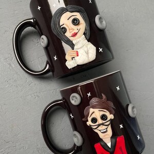 May include: Two black ceramic mugs with white stars and gray buttons. One mug features a sculpted woman with black hair, a white sweater, and button eyes. The other mug features a sculpted man with brown hair, a red jacket, and button eyes.