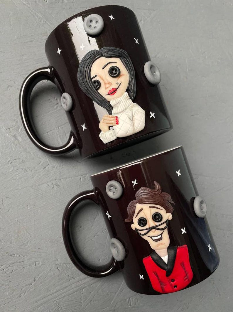 May include: Two black ceramic mugs with white stars and gray buttons. One mug features a sculpted woman with black hair, a white sweater, and button eyes. The other mug features a sculpted man with brown hair, a red jacket, and button eyes.