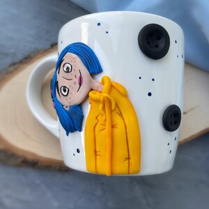 May include: White ceramic mug with a hand-painted illustration of a girl with blue hair wearing a yellow raincoat. The mug has black buttons on the side.
