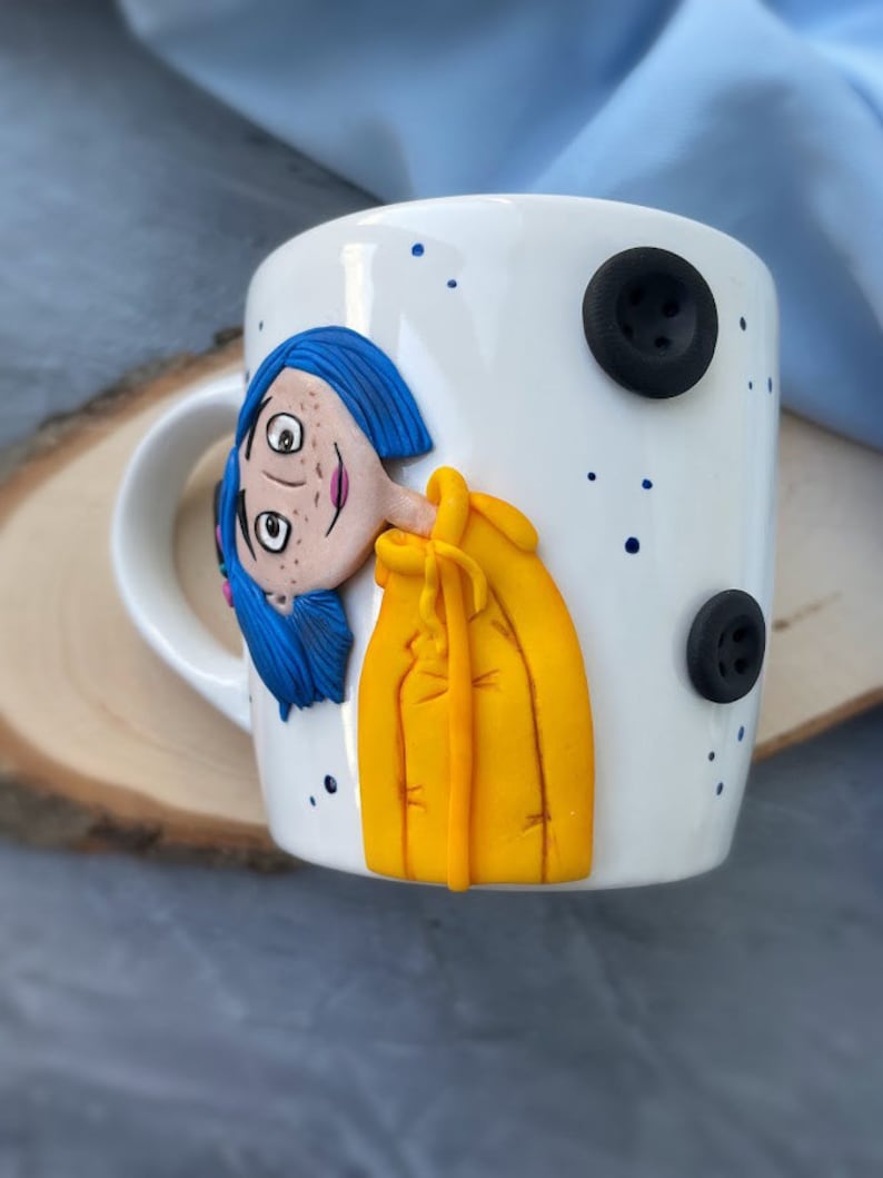May include: White ceramic mug with a hand-painted illustration of a girl with blue hair wearing a yellow raincoat. The mug has black buttons on the side.