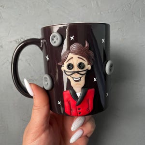 May include: A dark brown ceramic mug with a 3D sculpted image of a man with brown hair, a red jacket, and a white shirt. The mug has a white handle and is decorated with gray buttons.