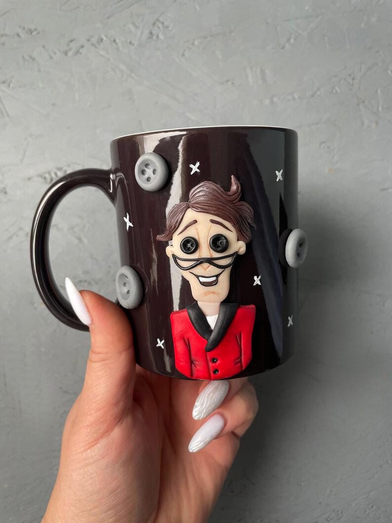 May include: A dark brown ceramic mug with a 3D sculpted image of a man with brown hair, a red jacket, and a white shirt. The mug has a white handle and is decorated with gray buttons.