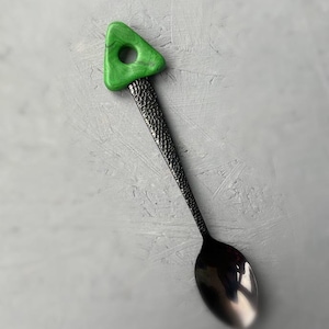 May include: A black metal spoon with a textured handle and a green triangle-shaped handle with a hole in the center.