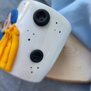 May include: White ceramic mug with a blue and yellow cartoon character on the front. The mug has two black buttons with four holes each.