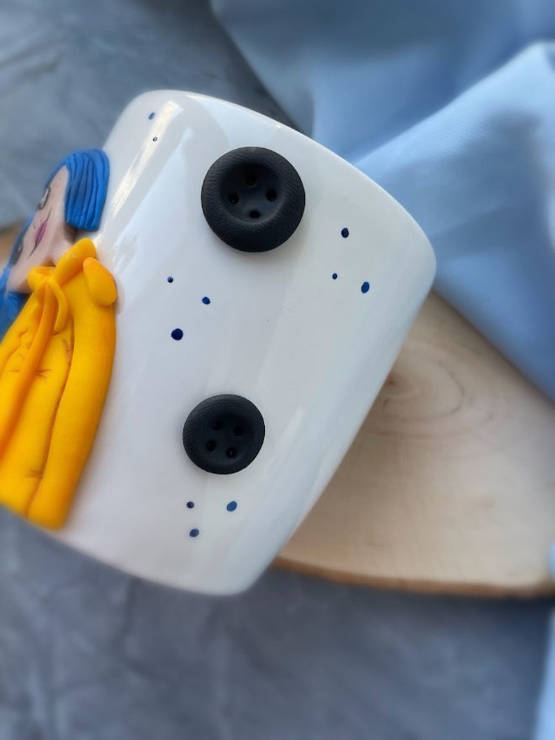 May include: White ceramic mug with a blue and yellow cartoon character on the front. The mug has two black buttons with four holes each.