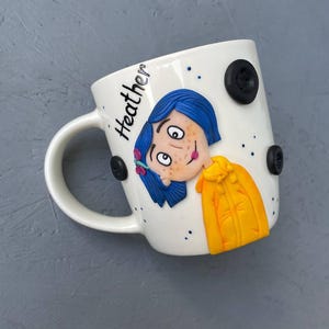May include: White ceramic mug with a hand-painted illustration of a girl with blue hair and a yellow jacket. The mug has black buttons and the name "Heather" written on the side.