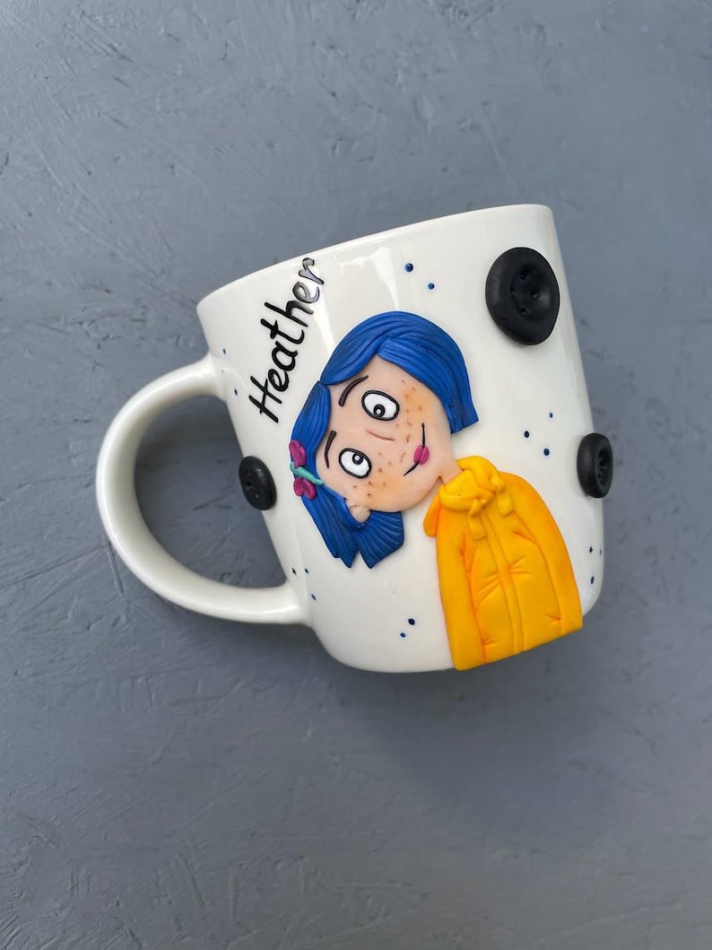 May include: White ceramic mug with a hand-painted illustration of a girl with blue hair and a yellow jacket. The mug has black buttons and the name "Heather" written on the side.