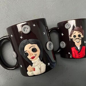 May include: Two black ceramic mugs with hand-painted details. One mug features a woman with black hair and a white sweater, the other mug features a man with brown hair wearing a red jacket. Both mugs have gray buttons and white X's on the surface.