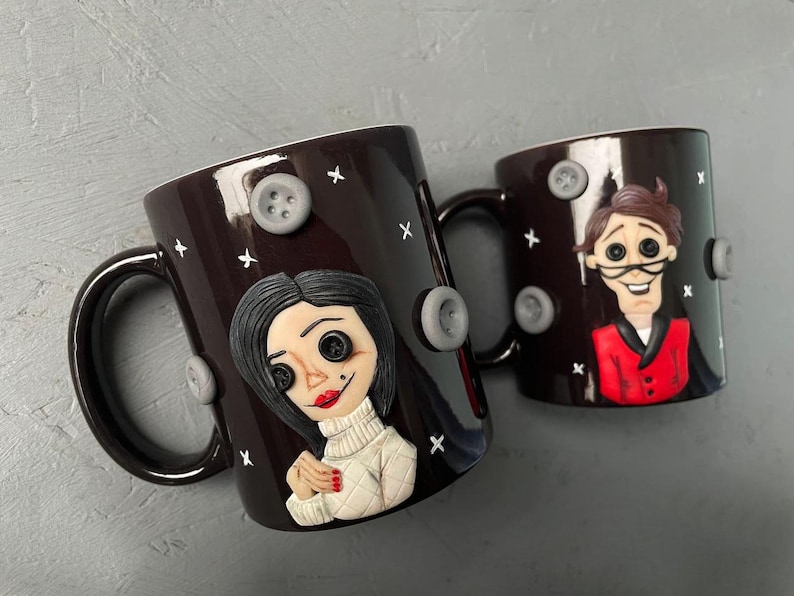 May include: Two black ceramic mugs with hand-painted details. One mug features a woman with black hair and a white sweater, the other mug features a man with brown hair wearing a red jacket. Both mugs have gray buttons and white X's on the surface.