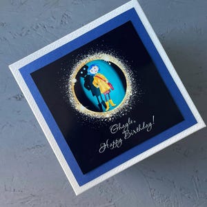 May include: A birthday card with a blue and white border. The card features a black and white image of a girl in a yellow raincoat standing in front of a blue background. The text "Hayle, Happy Birthday!" is written in white cursive on the card.