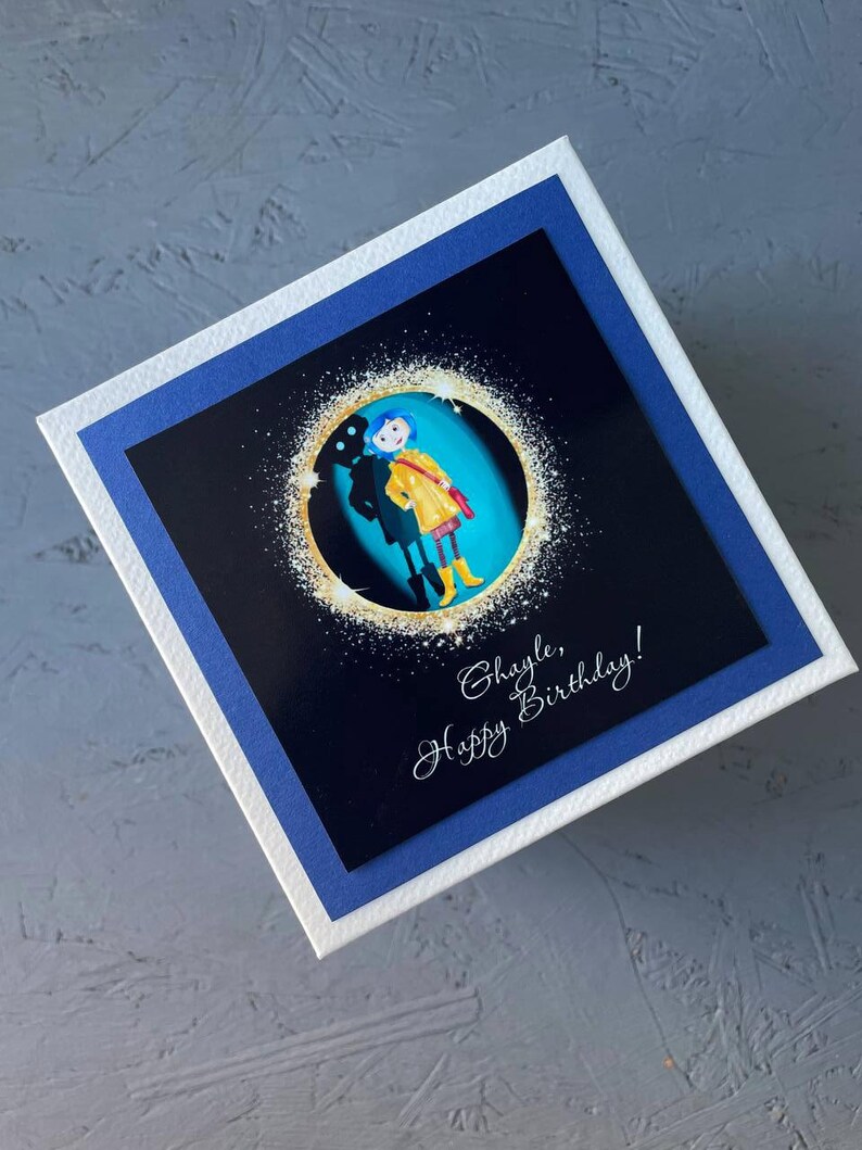 May include: A birthday card with a blue and white border. The card features a black and white image of a girl in a yellow raincoat standing in front of a blue background. The text "Hayle, Happy Birthday!" is written in white cursive on the card.