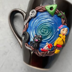 May include: Black ceramic mug with a blue and green swirl design. The mug has a 3D sculpted design featuring a black cat, a mouse wearing a red hat, and a girl with yellow hair and a blue dress.
