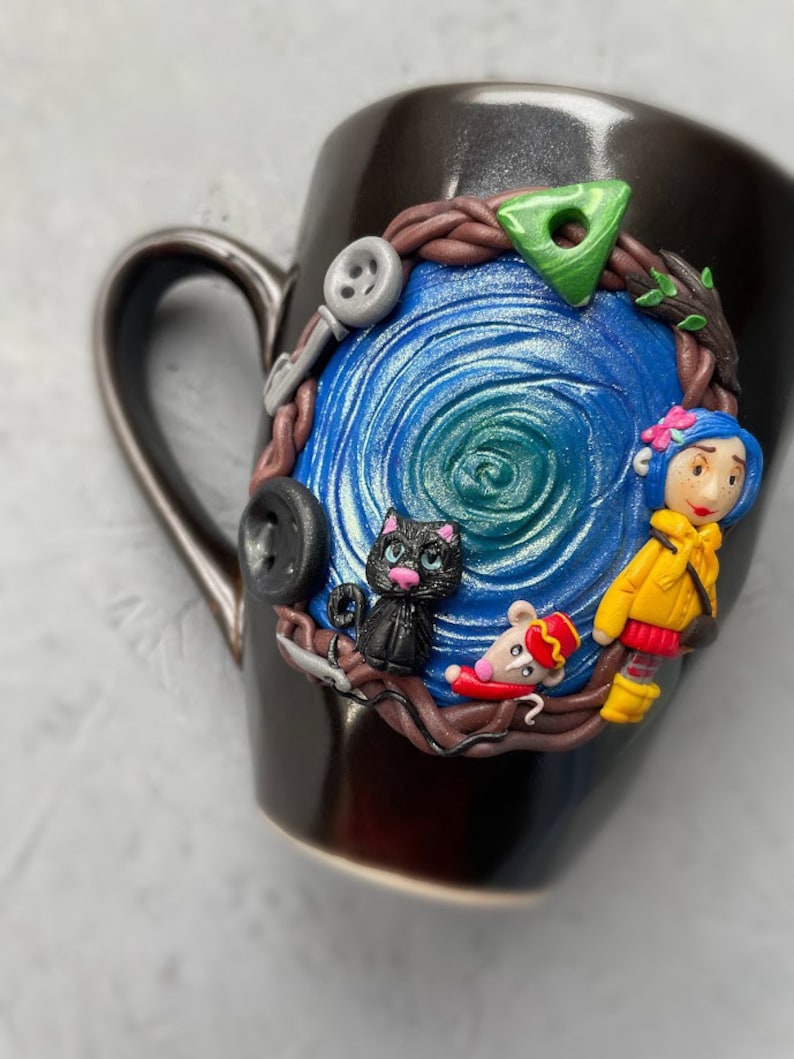 May include: Black ceramic mug with a blue and green swirl design. The mug has a 3D sculpted design featuring a black cat, a mouse wearing a red hat, and a girl with yellow hair and a blue dress.