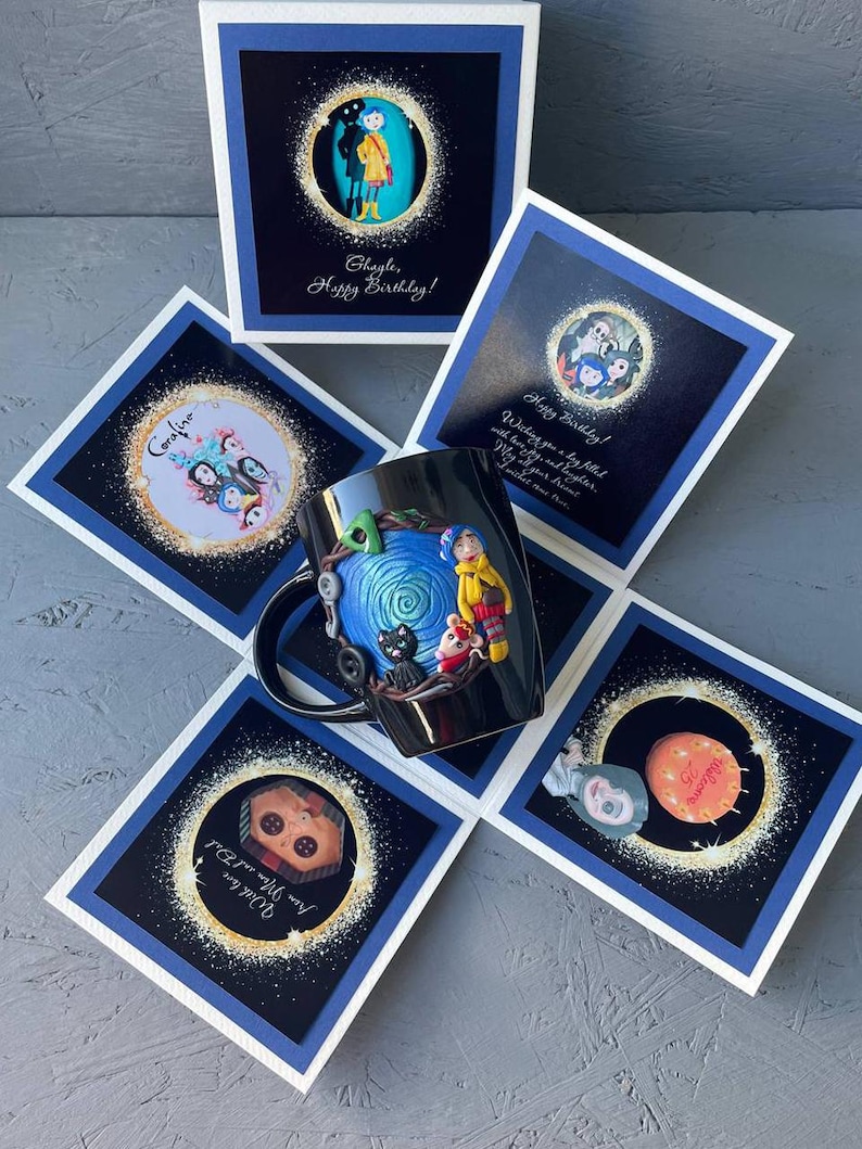May include: A black mug with a blue and white spiral design, a black cat, and a girl with red hair and a yellow jacket. The mug is surrounded by five white cards with blue borders. The cards have images of characters from the movie Coraline and the text "Coraline", "Ghagle, Happy Birthday!" and "Happy Birthday! Wishing you a big year with family and friends. May all your dreams come true."
