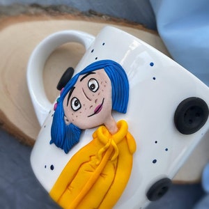 May include: White ceramic mug with a hand-painted image of a girl with blue hair wearing a yellow jacket. The mug has blue dots and black buttons.