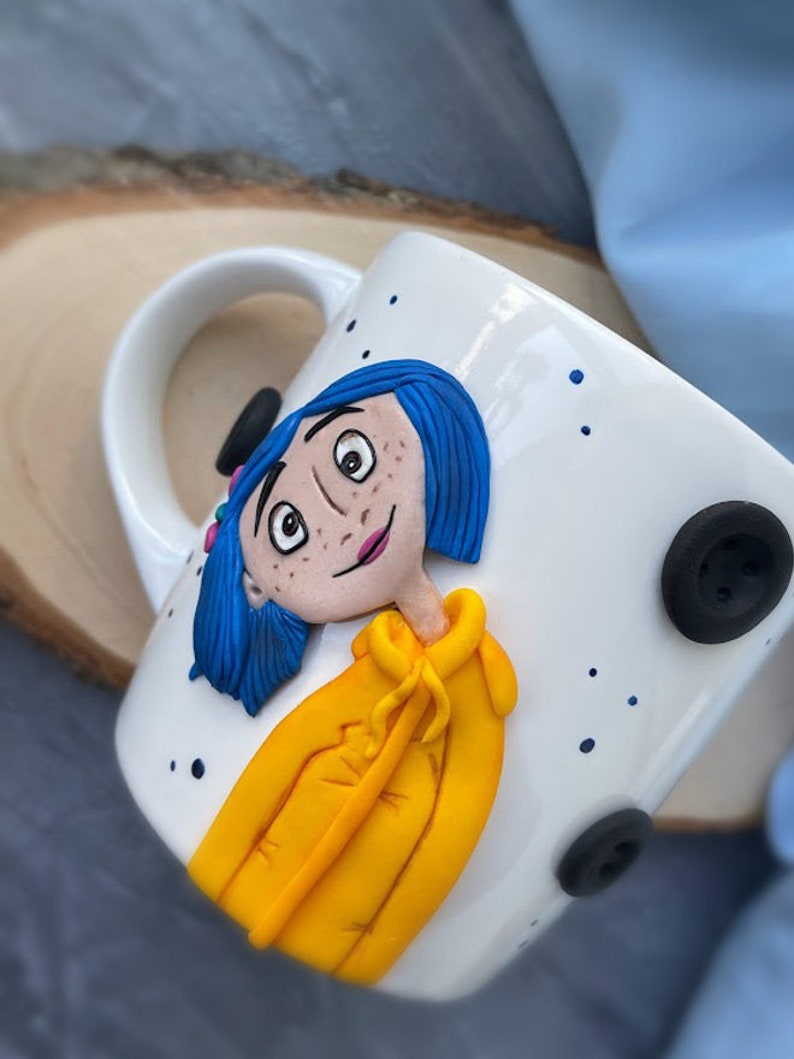 May include: White ceramic mug with a hand-painted image of a girl with blue hair wearing a yellow jacket. The mug has blue dots and black buttons.