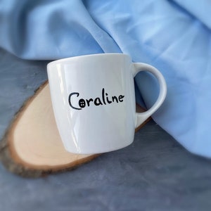 May include: White ceramic mug with a black handwritten name "Coraline" on the side.