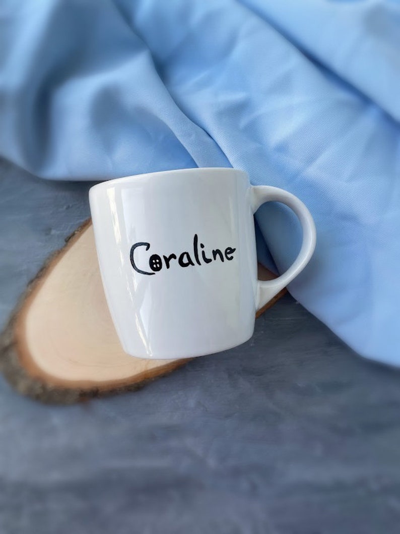 May include: White ceramic mug with a black handwritten name "Coraline" on the side.
