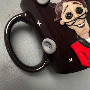 May include: A dark brown ceramic mug with a white and black illustration of a character with a red jacket and large button eyes. The mug has a white handle and is decorated with white stars and gray buttons.