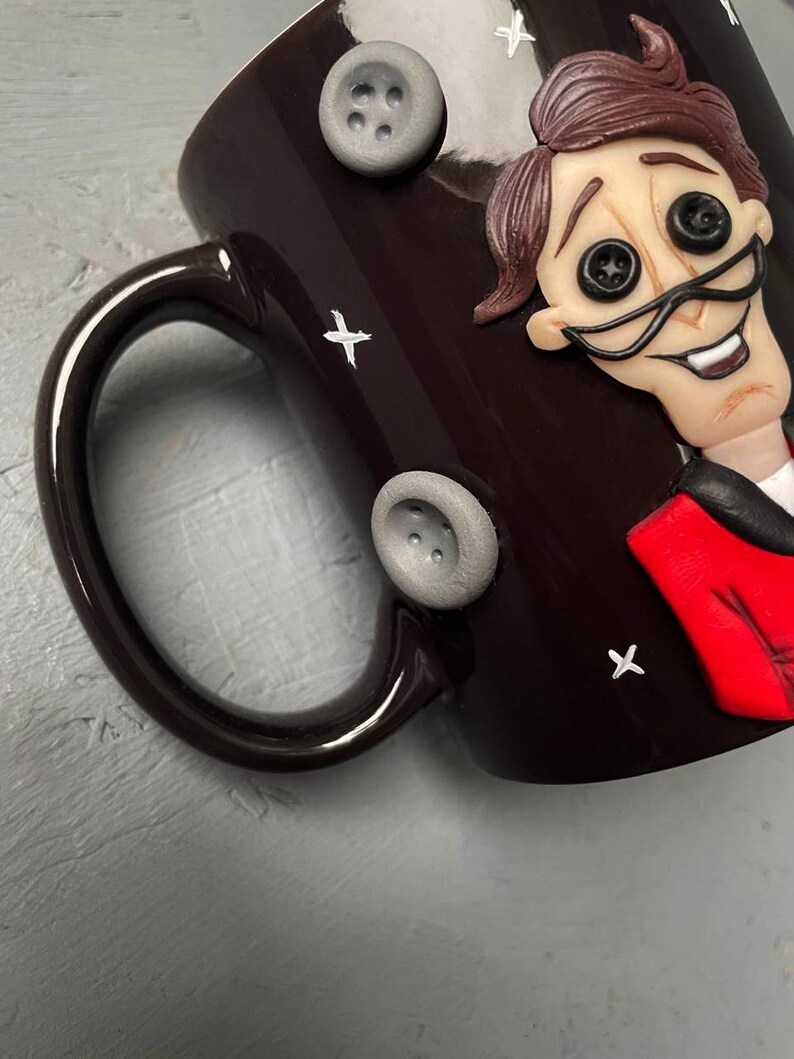 May include: A dark brown ceramic mug with a white and black illustration of a character with a red jacket and large button eyes. The mug has a white handle and is decorated with white stars and gray buttons.