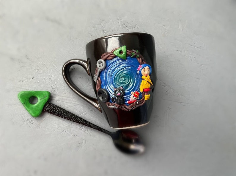 May include: Black ceramic mug with a hand-painted design featuring a girl with a yellow jacket, a black cat, and a mouse. The design is surrounded by a blue and green swirl and brown vines.