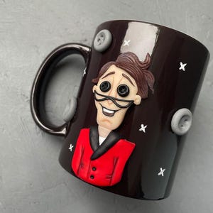 May include: A dark brown ceramic mug with a white glaze. The mug features a 3D sculpted image of a man with brown hair, a red jacket, and a black bow tie. The man has a large smile and large black eyes. The mug is decorated with white stars and gray buttons.