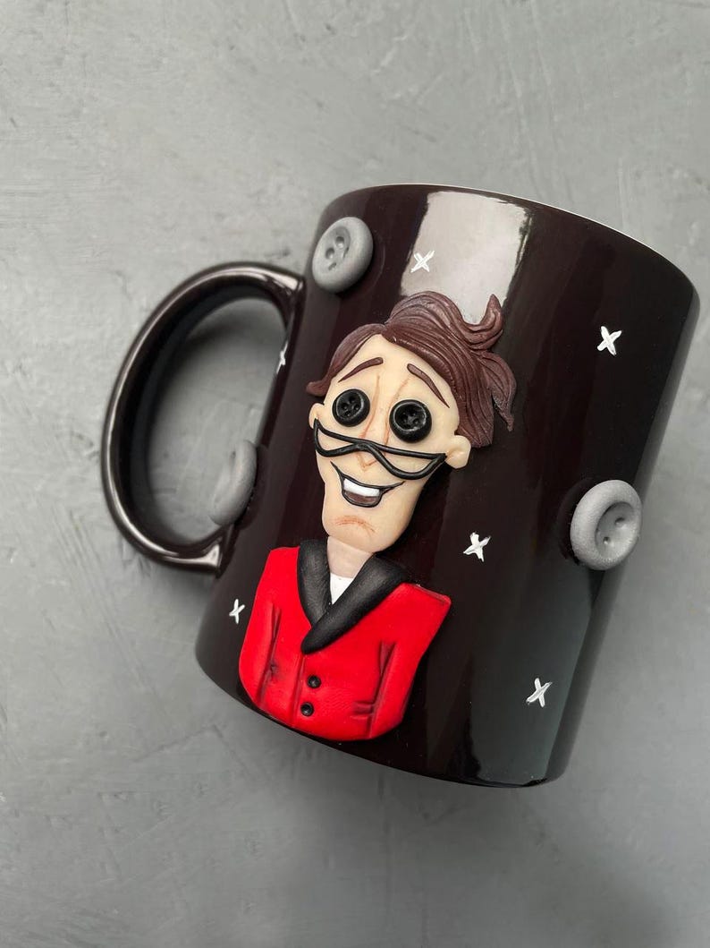 May include: A dark brown ceramic mug with a white glaze. The mug features a 3D sculpted image of a man with brown hair, a red jacket, and a black bow tie. The man has a large smile and large black eyes. The mug is decorated with white stars and gray buttons.