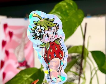May include: A holographic sticker featuring a cartoon strawberry fairy with a green leafy crown and a white flower. The fairy is wearing a red strawberry costume with green leaves and white seeds.