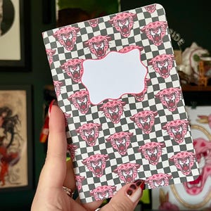 May include: A pink and black chequered notebook with a pink panther face pattern. The notebook has a blank white label with a wavy border.