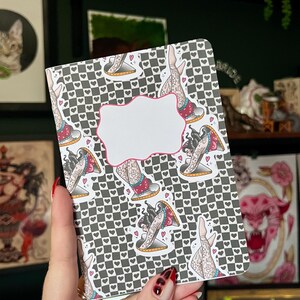 May include: A small black and white chequered notebook with a repeated pattern of a woman with tattoos in a red and white dress. The notebook has a blank white label on the front.