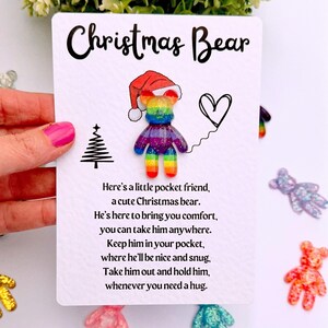 May include: A white card with the text "Christmas Bear" and a rainbow-coloured bear wearing a Santa hat. The card includes a message about a pocket-sized Christmas bear for comfort and hugs. Small bear-shaped decorations are scattered around.