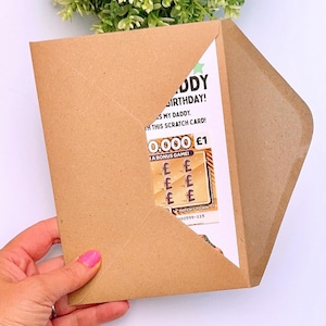 Puede incluir: Un sobre de papel marr&oacute;n con una tarjeta de cumplea&ntilde;os. La tarjeta dice "&iexcl;Feliz Cumplea&ntilde;os Pap&aacute;!" y presenta un juego de rascar con la posibilidad de ganar &pound;1. El sobre est&aacute; sostenido por una mano.