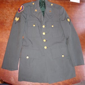 May include: An olive green military jacket with gold-colored buttons and insignia. The jacket has multiple pockets and patches on the sleeves. The interior lining is a darker green. A classic piece of military apparel.