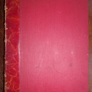 May include: A vintage book with a red cover and spine. The spine is a darker shade of red with a textured, leather-like appearance. The cover is a solid, lighter red with a subtle vertical texture.