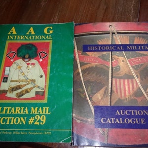 May include: Two auction catalogs for historical militaria. The first catalog is green with a white border and features a man in a white uniform with a fur hat. The second catalog is red with a blue border and features a drum with an eagle and American flag design. The text on the first catalog reads "AAG International Militaria Mail Auction #29". The text on the second catalog reads "Historical Militaria Auction Catalogue 130A".