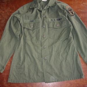 May include: Olive green military shirt with long sleeves, two chest pockets, and button-down closure. Features the text "FAT" above the left pocket and "U.S. ARMY" above the right pocket. A patch is visible on the left sleeve.